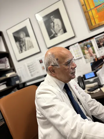 Photo of Dr. Raza seated at the desk of his office, attention towards his computer off-camera. Behind him on the wall are framed portraits of two of his mentors.