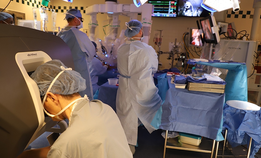 Photograph showing a surgeon in the foreground working at a da Vinci machine while other surgeons are in the background with the patent and robotic arms.