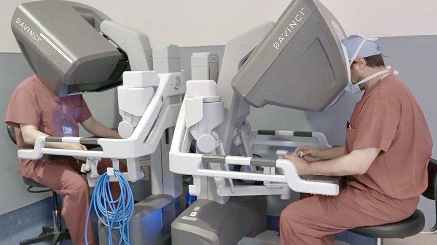 two surgeons sit and side by side robotic surgery consoles in the operating room.