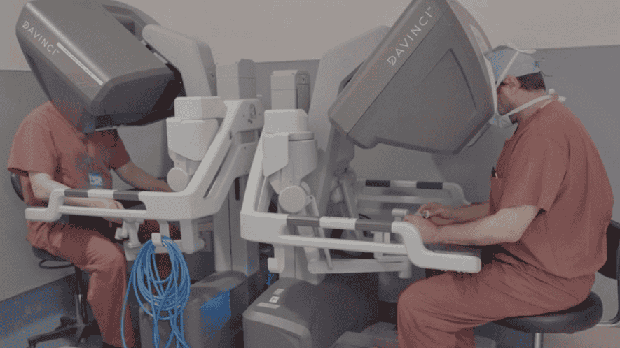 Two doctors seated at robotic machines looking into them and using hand controls