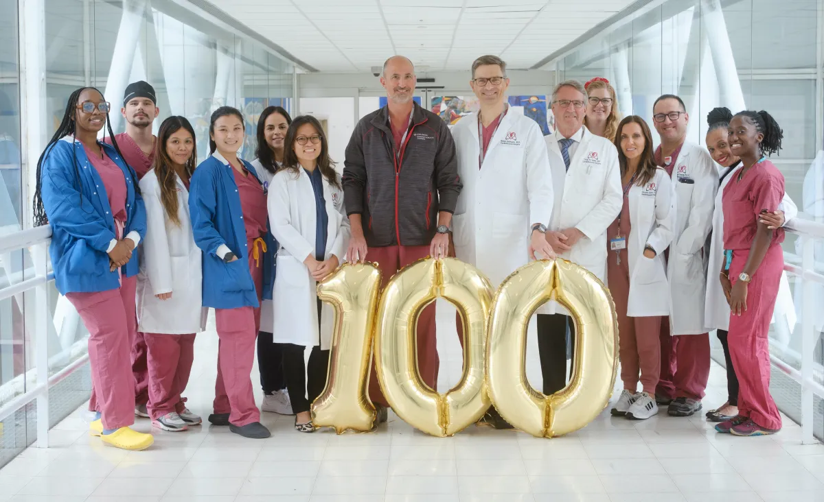 Robotic heart surgery team stands together with balloons that display "100" on hospital skybridge.
