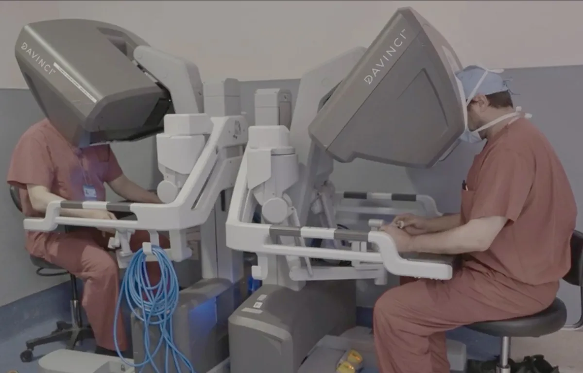 two surgeons sit and side by side robotic surgery consoles in the operating room.