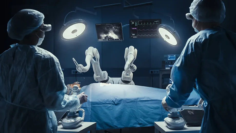 Photograph of two surgeons using robots controls as they look at an operating table with robotic arms that hover over a patient who is on the operating table.