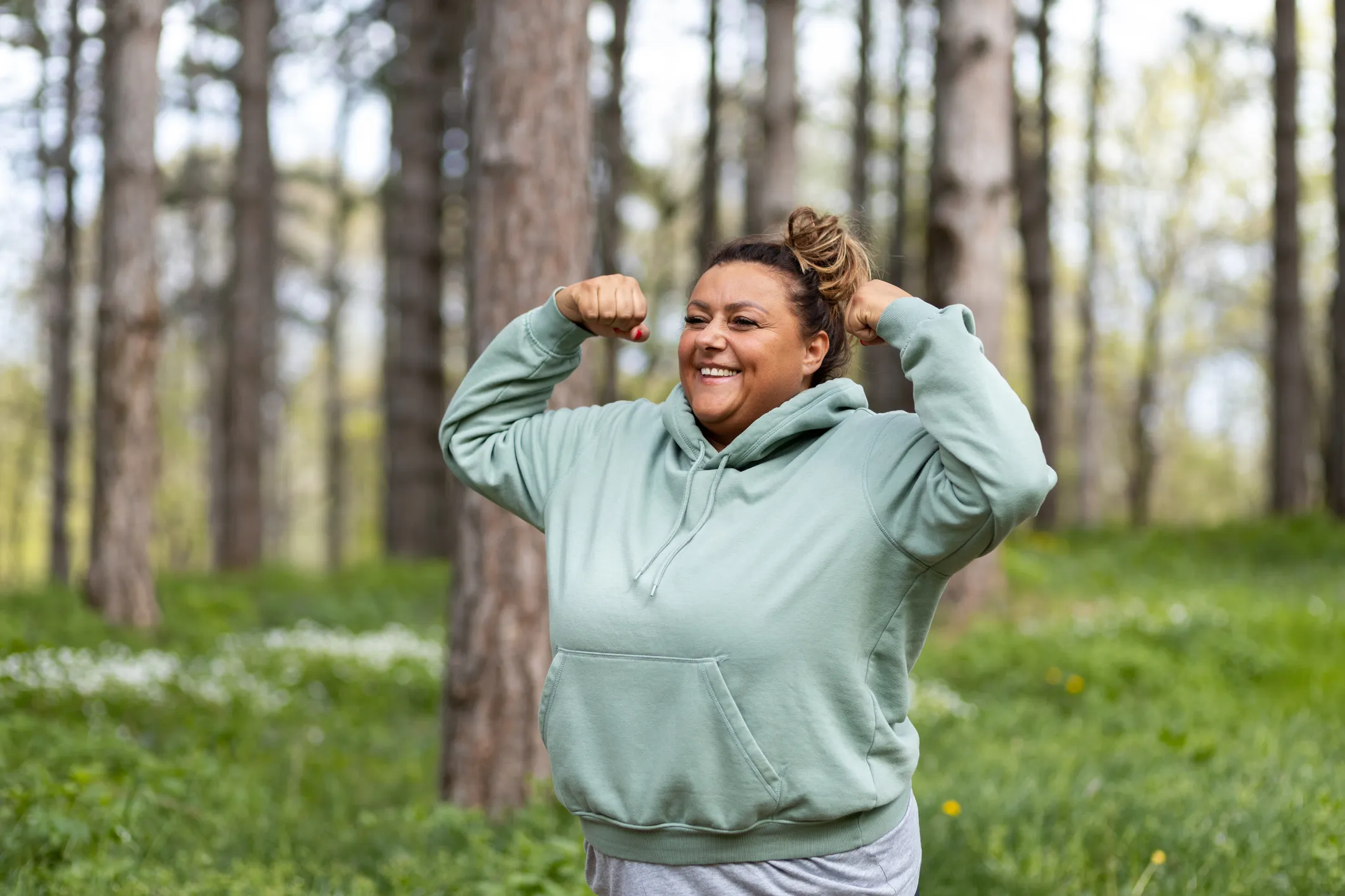 A woman proudly flexes while enjoying the outdoors in a woodsy setting.