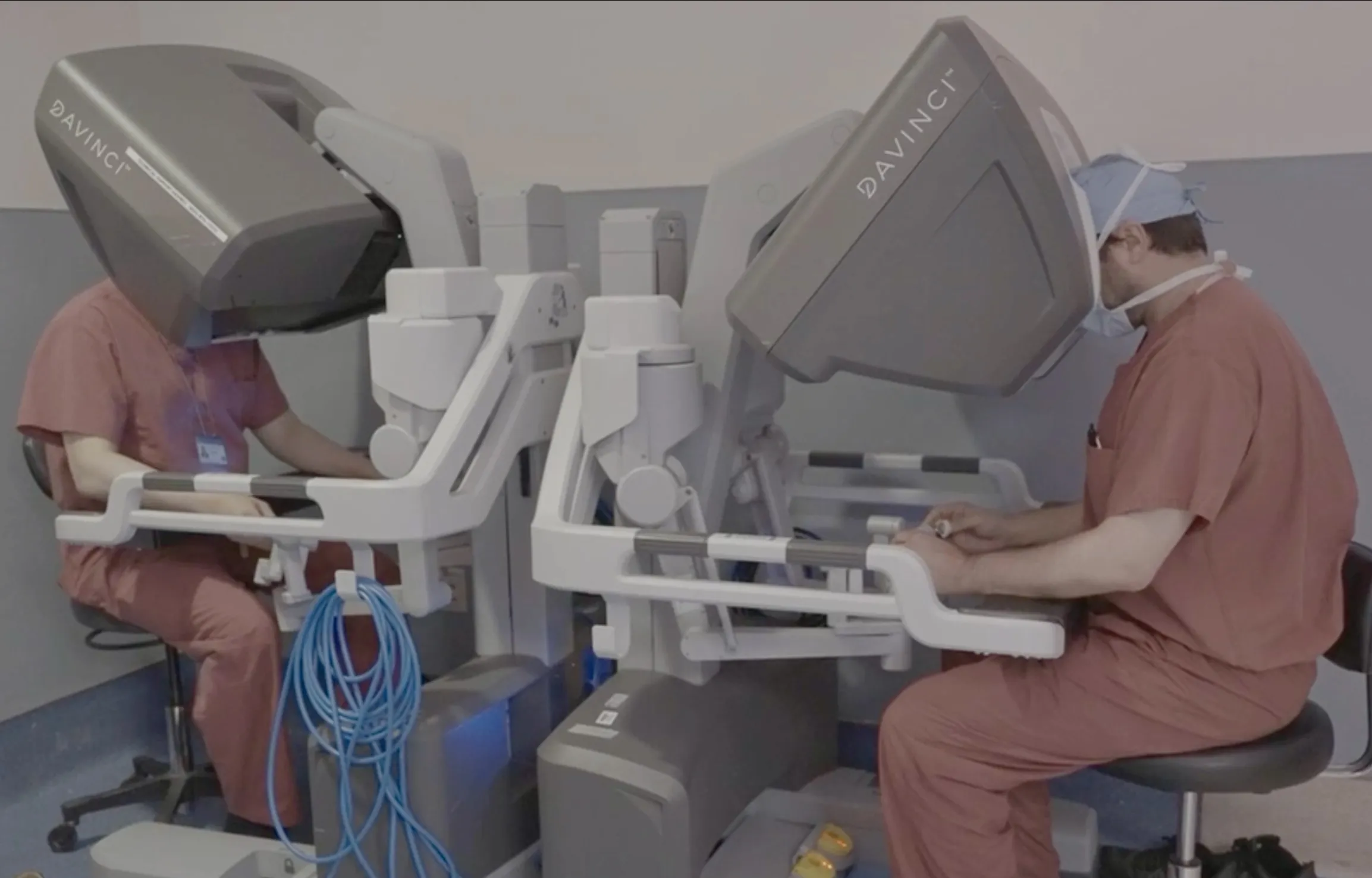 two surgeons sit and side by side robotic surgery consoles in the operating room.