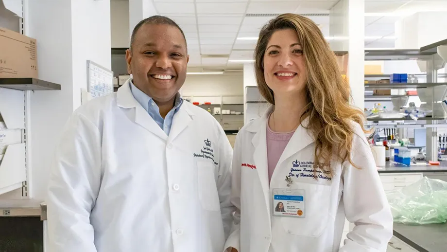 Photograph of Doctors Joel Gabre and Yoanna Pumpalova standing next to one another in a lab dressed in white lab coats.